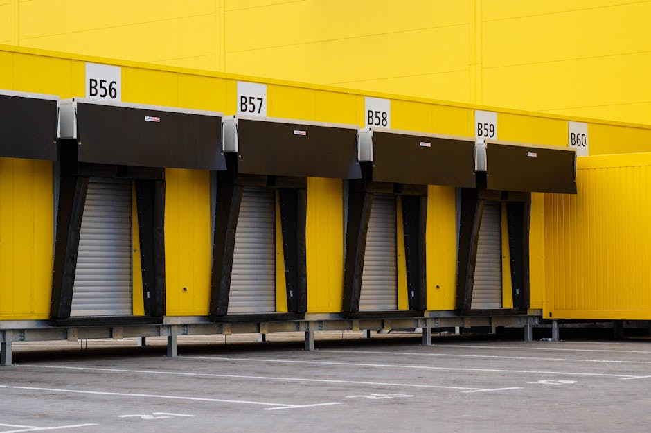 This image shows a row of four yellow loading bays at a warehouse or retail store, each equipped with black retractable dock shelters for moving trucks. The bays are numbered B56, B57, B58, and B59, with small white signage displaying the numbers above each dock. The dock shelters are partially open, revealing metallic roller shutters behind them. The area in front of the loading bays is an empty parking space with painted white lines for vehicle positioning. The scene appears to be outdoors, under natural lighting, with the yellow building’s exterior wall extending across the background. This setting is typical for furniture transport, home relocation, and moving logistics operations, where items are loaded or unloaded using trolleys or hand carts, often associated with professional removals such as those provided by Man with Van Bellingham.