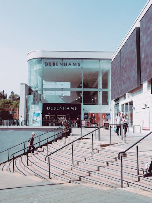 A modern, curved glass-fronted building with the name 'Debenhams' displayed above the entrance, situated at the top of wide outdoor steps with black metal handrails. The steps lead up from a paved surrounding area where several pedestrians are walking, some carrying shopping bags or backpacks. The building is part of Bellingham Shopping Centre, which features contemporary architectural design with a mix of glass and dark paneling. The sky is clear and blue, and local greenery can be seen in the background. The scene depicts a busy retail environment, consistent with a typical shopping centre setting, which may be relevant to house removals or relocation services involving shopping complexes. Man with Van Bellingham offers removals services, including moving household or retail goods, in locations like this. The image naturally supports topics related to moving logistics, transportation, and packing during home or commercial relocations.