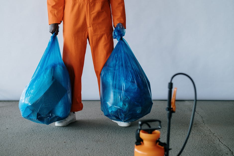 A person wearing orange work trousers and white shoes is standing indoors on a concrete floor, holding two large blue plastic bags filled with waste materials, positioned near the lower part of their body. The person's upper body and head are not visible in the image. The background features a plain, light-colored wall. An orange and black backpack sprayer with a hose and spray nozzle is placed on the floor in the foreground, slightly out of focus. The scene depicts a waste removal process related to home relocation or packing and moving activities, with the individual preparing bulky waste for collection or disposal, consistent with services offered by Man with Van Bellingham in the realm of removals and waste management.