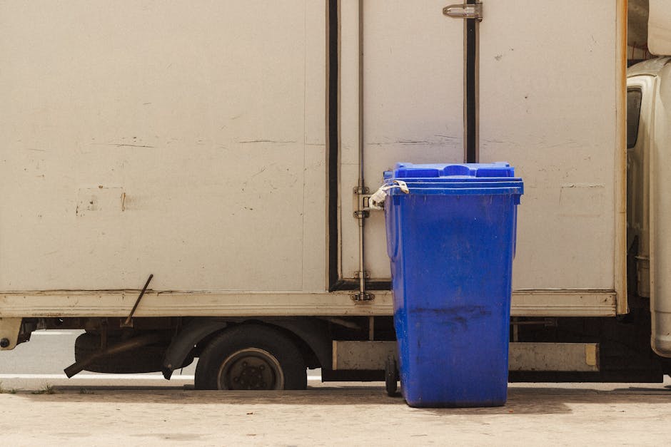 A blue wheeled waste bin positioned directly in front of a white delivery or storage truck parked outside a residential property. The bin is made of plastic and has a closed lid, situated on a paved surface next to the truck's rear loading area. The truck's side panel appears somewhat dirty and weathered, with visible marks and grime. Part of a driveway or street is visible at the bottom of the image, with a small patch of grass near the vehicle's wheel. The setting suggests an accessible location for loading or collection, possibly related to home removal or waste disposal services conducted by Man with Van Bellingham, as indicated by the context of the page about bulky waste pickup versus council collection, focusing on moving logistics and clearance tasks.