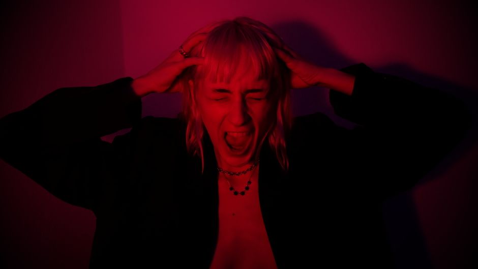 A woman with blonde hair and wearing dark clothing is inside a room, standing near a doorway, with her hands placed on her head and her mouth open in an expression of frustration or stress. The scene is illuminated with red lighting, creating a tense atmosphere. Visible behind her are some cardboard boxes and wrapped furniture, suggesting a home relocation or moving process. The boxes are stacked on the floor, and some items are covered with protective fabric or plastic wrap. The doorway behind her leads to another part of the property, and the overall environment appears to be in the midst of packing and loading for a move, reflecting the challenges associated with furniture transport and moving logistics. The scene aligns with themes of house removals and professional moving services, such as those offered by Man with Van Bellingham.