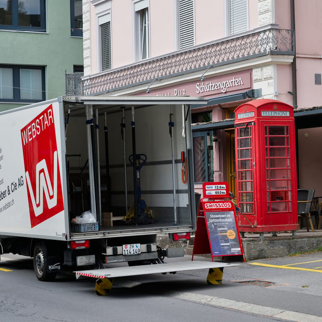A small white relocation van with the logo 'WEBSTAR' parked on a city street in front of a pink and white building with a balcony and decorative railing. The back of the van is open, revealing interior equipment such as a hand truck, straps, and packing materials, indicating a home relocation or furniture transport process. The van is positioned on a loading ramp with wheel chocks for stability. Nearby, a traditional red British telephone box stands beside a sidewalk with yellow parking bay markings, and an outdoor cafe area with wooden tables and chairs. The environment appears urban, with natural daylight illuminating the scene, capturing the practical aspects of moving and packing services in preparation for a house removal or transfer of goods, as offered by Man with Van Bellingham.