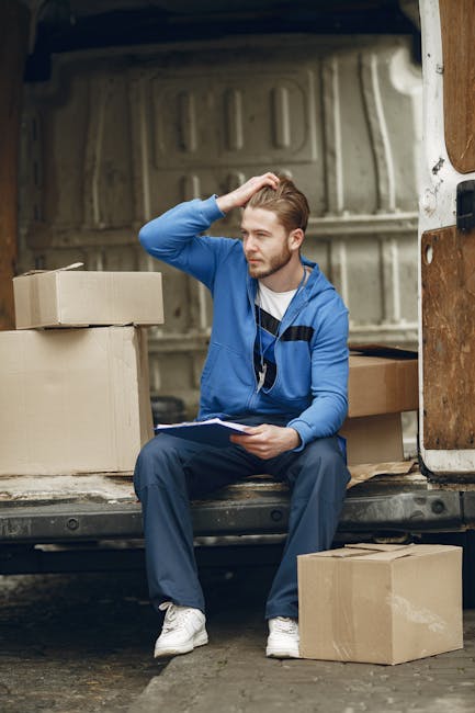 A woman with blonde hair and wearing dark clothing is inside a room, standing near a doorway, with her hands placed on her head and her mouth open in an expression of frustration or stress. The scene is illuminated with red lighting, creating a tense atmosphere. Visible behind her are some cardboard boxes and wrapped furniture, suggesting a home relocation or moving process. The boxes are stacked on the floor, and some items are covered with protective fabric or plastic wrap. The doorway behind her leads to another part of the property, and the overall environment appears to be in the midst of packing and loading for a move, reflecting the challenges associated with furniture transport and moving logistics. The scene aligns with themes of house removals and professional moving services, such as those offered by Man with Van Bellingham.