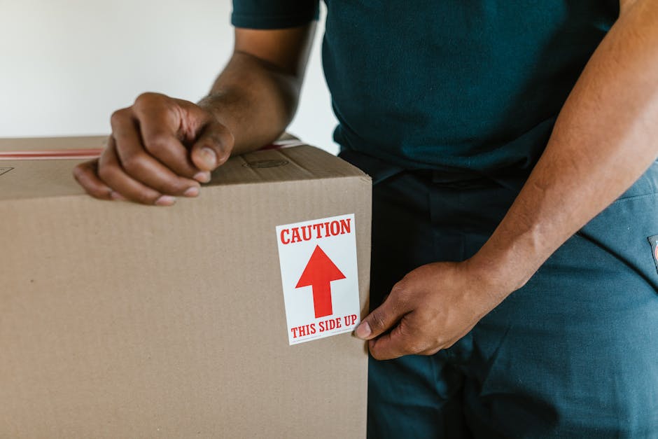 A person wearing a dark green T-shirt and dark trousers is handling a cardboard moving box, which has a red and white caution label affixed to its side indicating 'This Side Up' with a downward arrow.