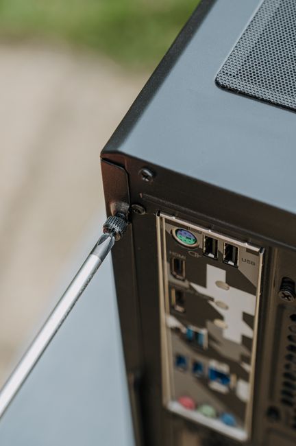 Close-up image of the rear panel of a black computer tower showing various input and output ports, including USB ports, audio jacks, and expansion slots, with a screwdriver inserted into a screw at th