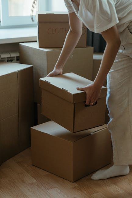 A person in light-colored clothing is in an indoor space, lifting and stacking three cardboard boxes among other packed moving cartons. The boxes vary in size, with the smallest being held securely, and the larger ones placed on the floor or on top of each other. The boxes are sealed with brown packing tape; one has the word 'CLOTHES' written on it in red marker. The background shows a window with natural light illuminating the room, which has a wooden floor. This scene depicts the packing and home relocation process, where items are being prepared for moving, an activity often handled by professional removals services like Man with Van Bellingham. The arrangement of boxes suggests an organized packing effort in readiness for furniture transport and the logistical steps involved in a house move.