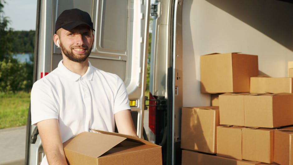 A young man with a beard, wearing a black cap and a white polo shirt, is standing outside during daylight, holding a medium-sized cardboard box. He is positioned next to an open delivery van with its rear doors wide open, revealing several stacked cardboard boxes inside and on a nearby pavement area. The van is parked on a paved surface, with green trees and a body of water visible in the background, indicating an outdoor setting. The scene depicts a home relocation or furniture transport process, with the man preparing to load or unload boxes, supported by a trolley and packing materials visible in the environment. This visual aligns with professional removal services such as those offered by Man with Van Bellingham, emphasizing the logistics involved in quick and efficient last-minute moves in SE6.