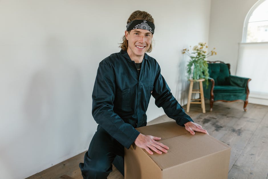 A person in light-colored clothing is in an indoor space, lifting and stacking three cardboard boxes among other packed moving cartons. The boxes vary in size, with the smallest being held securely, and the larger ones placed on the floor or on top of each other. The boxes are sealed with brown packing tape; one has the word 'CLOTHES' written on it in red marker. The background shows a window with natural light illuminating the room, which has a wooden floor. This scene depicts the packing and home relocation process, where items are being prepared for moving, an activity often handled by professional removals services like Man with Van Bellingham. The arrangement of boxes suggests an organized packing effort in readiness for furniture transport and the logistical steps involved in a house move.