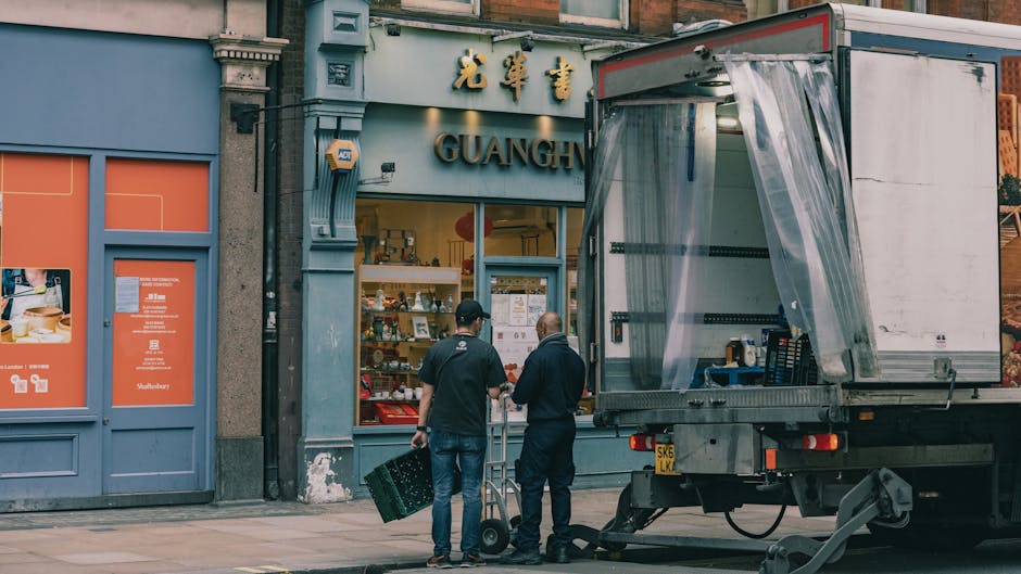 Two removal workers are preparing to load a moving truck parked on the pavement outside a storefront with Chinese characters and the word 'GUANG' visible above the entrance. One worker, wearing a black uniform and cap, is carrying a small black crate while the other, dressed in a dark jacket, stands next to a hand truck or dolly. The open truck has a plastic sheet hanging at the side, revealing partially visible packaging supplies and equipment inside. Several cardboard boxes, some wrapped in plastic, are positioned on the pavement near the truck, suggesting a home relocation or furniture transport operation. The storefront features large windows and painted walls, indicating an urban shopping district, and the scene is lit by natural daylight. This image is associated with professional removals services, such as those offered by Man with Van Bellingham, and showcases the packing and loading process integral to house removals and moving logistics.