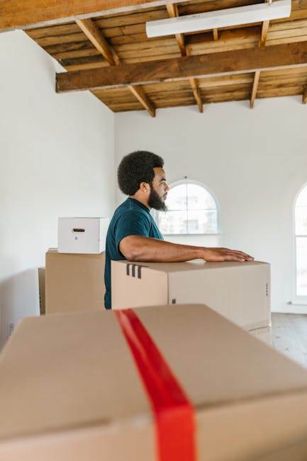 A man with dark curly hair and a beard, wearing a blue shirt, is inside a spacious, well-lit room with white walls and a wooden ceiling structure. He is gently resting his hands on top of cardboard boxes stacked along the floor, indicating an ongoing home relocation or packing process. Several cardboard boxes of various sizes, some sealed with packing tape and others with red strapping or reinforced corners, are arranged around him, suggesting careful packing for moving. The room features two arched windows that allow natural light to illuminate the scene, and the background shows a clean, minimally furnished interior. This setting reflects a residential environment in the midst of furniture transport and packing, exemplifying professional removal services that facilitate home removals, with [COMPANY_NAME] potentially involved in the loading process. The overall scene captures a peaceful moment in a furniture transport and packing operation, supporting the topic of house removals and local moving tips.