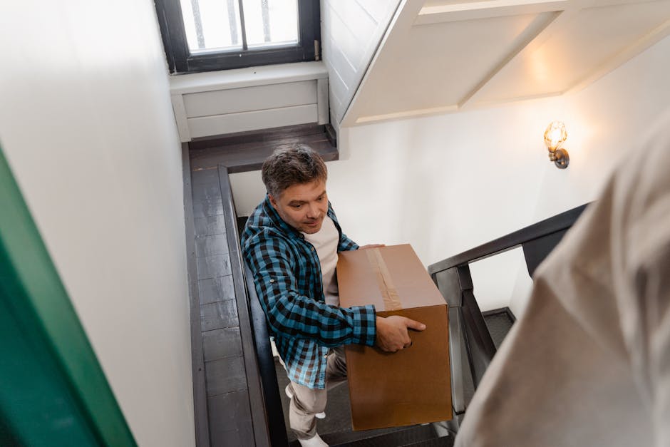 A man with short brown hair, dressed in a blue and black checkered shirt over a white undershirt, is carrying a medium-sized cardboard box while ascending a narrow staircase inside a home. The staircase has dark wooden steps with a black railing, and the walls are painted white. A small window at the top of the stairs allows natural light to illuminate the scene, complemented by a wall-mounted light fixture. The packing materials, including the cardboard box, are part of the home relocation process managed by Man with Van Bellingham, who specializes in removals and furniture transport, ensuring safe handling during the staircase move in Bellingham.