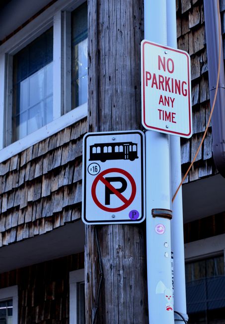 A photograph showing two traffic signs mounted on a weathered wooden utility pole next to a residential building with window frames and wooden siding. The upper sign is rectangular with a white background and red text stating 'NO PARKING ANY TIME,' indicating parking restrictions in the area. The lower sign is square with a white background, displaying a red circle with a diagonal line crossing out a black letter 'P', symbolising no parking, and an additional black icon of a bus and a trolleybus, suggesting restrictions for certain vehicles. The pole also has some small stickers and markings, and a set of orange wires runs vertically along it. The background includes part of a house with large windows and a clear, daytime sky, contextualising the scene as part of a street environment possibly linked to house removals or moving logistics, as referenced in the website page about parking suspension rules in SE6.