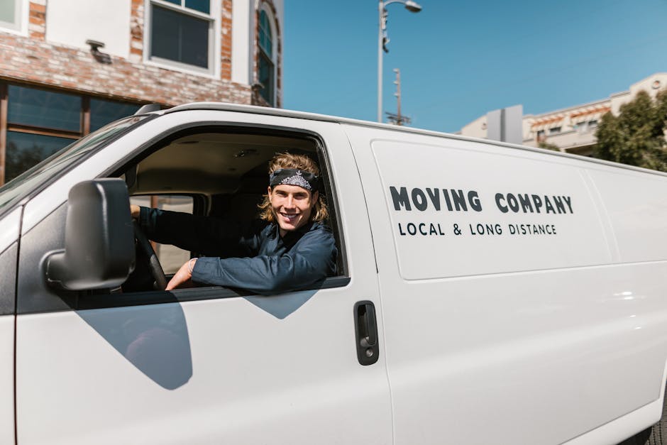 A man with long, light brown hair wearing a black bandana and a dark blue shirt is sitting in the driver's seat of a white moving van, smiling at the camera. The van has the words 'MOVING COMPANY LOCA