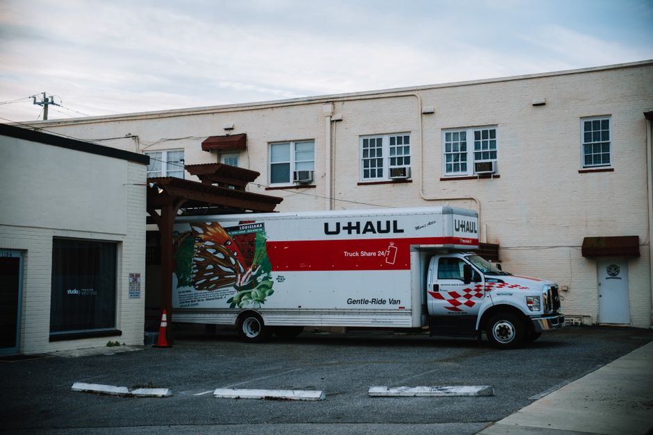 A man with long, light brown hair wearing a black bandana and a dark blue shirt is sitting in the driver's seat of a white moving van, smiling at the camera. The van has the words 'MOVING COMPANY LOCA